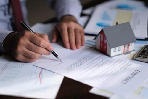 man signing papers scattered over a desk with small house model - update my estate plan concept