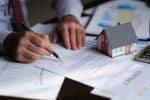 man signing papers scattered over a desk with small house model - update my estate plan concept